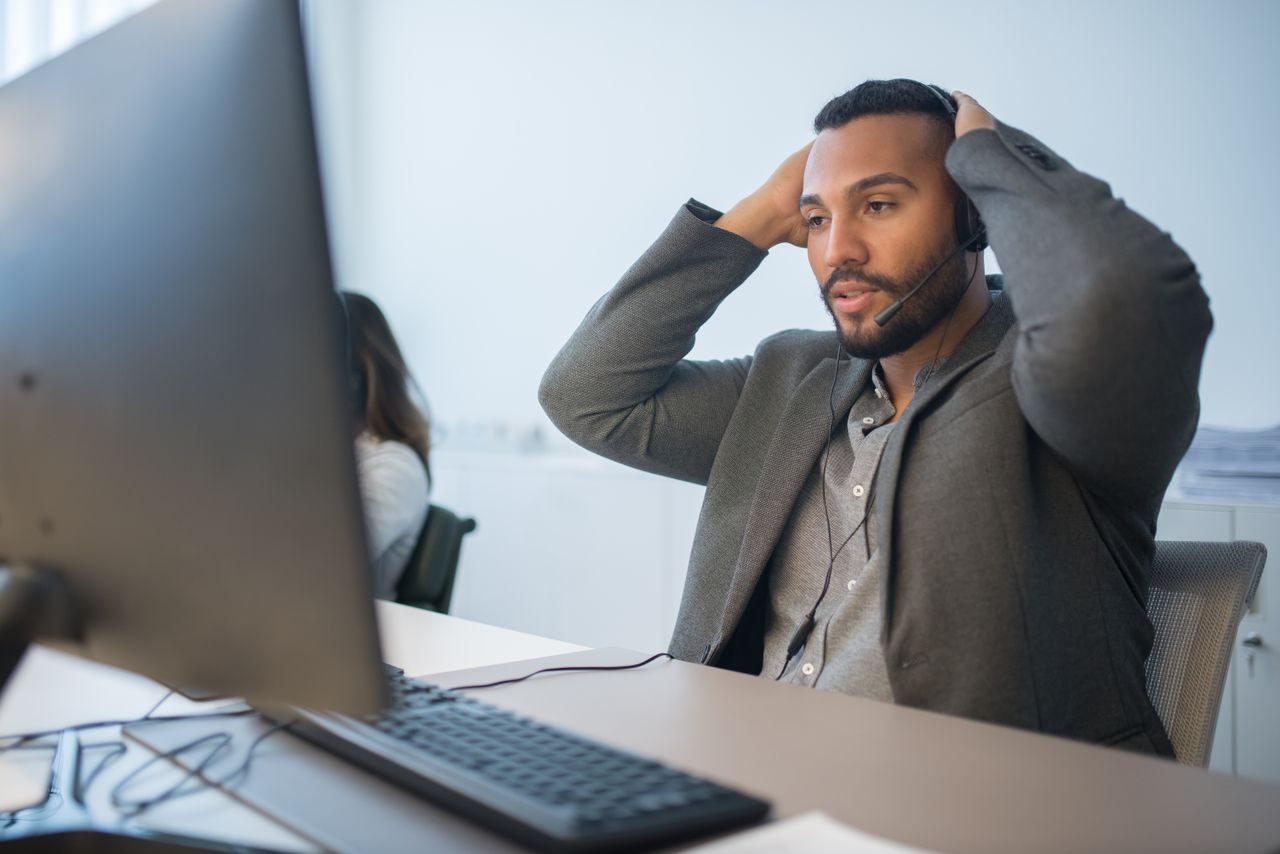 Man in a headset working at a desk, hands on head, conveys work stress.
